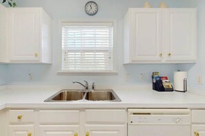 A kitchen with white cabinets, a double sink, a clock, and a window with blinds. - Ironwood - Turtle Beach (North Myrtle Beach)