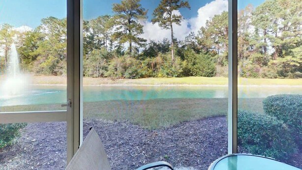 Ein Zimmer mit großem Fenster mit Blick auf einen See, Bäume und eine Fontäne.