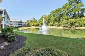 A landscaped pond with a fountain, surrounded by manicured grass and trees, with a building in the background. - Ironwood - Turtle Beach (North Myrtle Beach)