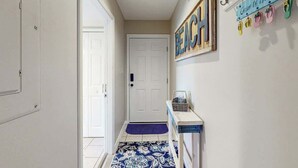 A hallway with a white door, a blue and white patterned rug, a small table with a basket, and a wall sign that reads 'BEACH'. - Live in the Sun, Play in the Sea (Surfside Beach)