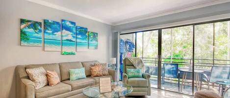 A living room with a sofa, a glass coffee table, and a view of the outdoors through large windows.