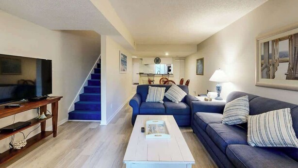 A living room with a TV, sofa, coffee table, and staircase leading to a kitchen. - Summertree Village A-11 (Myrtle Beach)