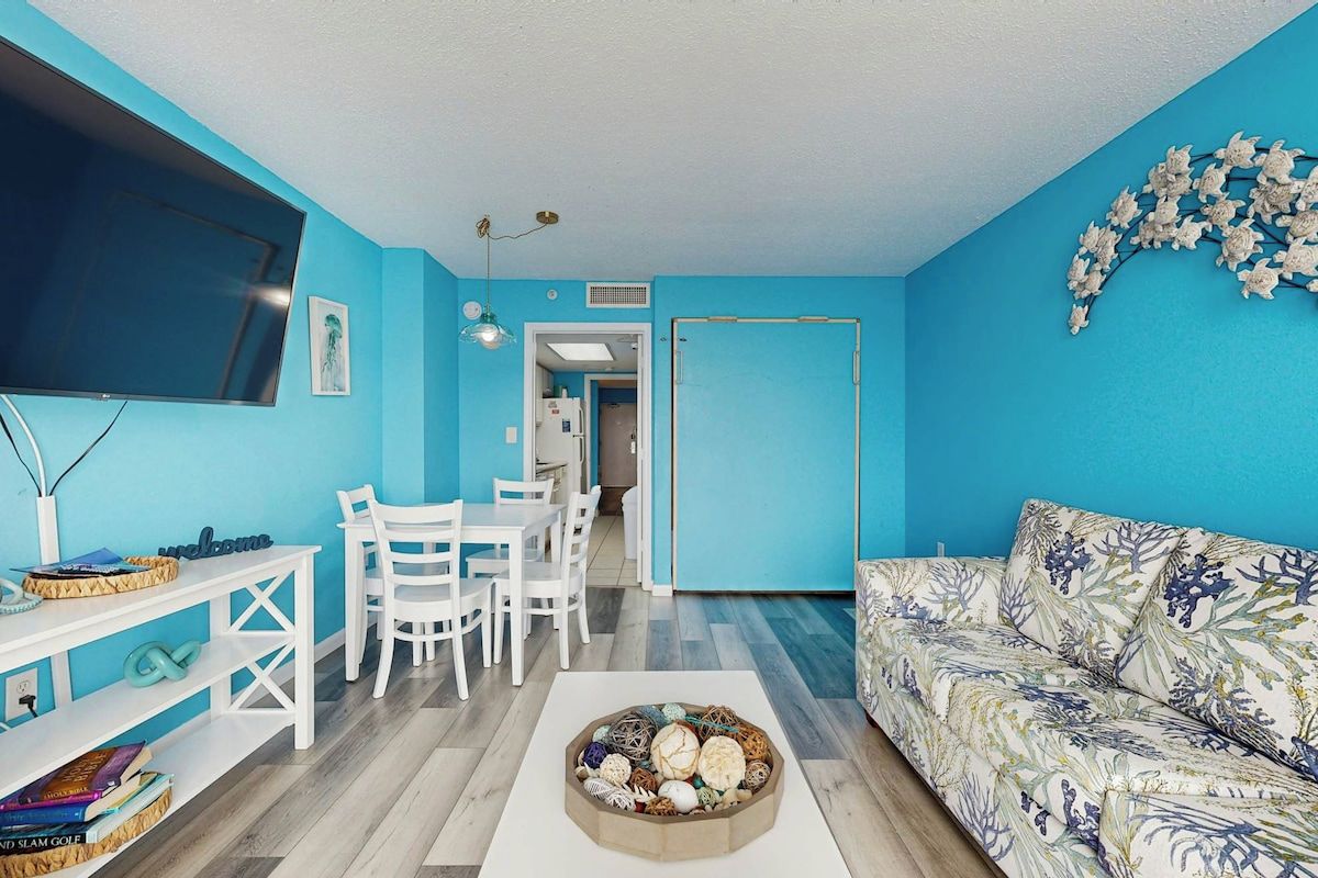 Una sala de estar con una pared azul, un televisor de pantalla plana, un sofá blanco, una mesa de centro con objetos decorativos y una zona de comedor con sillas y una mesa blancos.