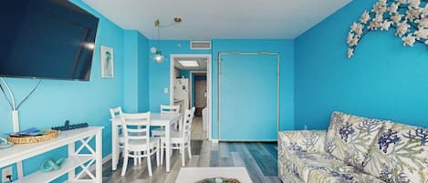 A living room with a blue wall, a flat-screen TV, a white sofa, a coffee table with decorative items, and a dining area with white chairs and a table.