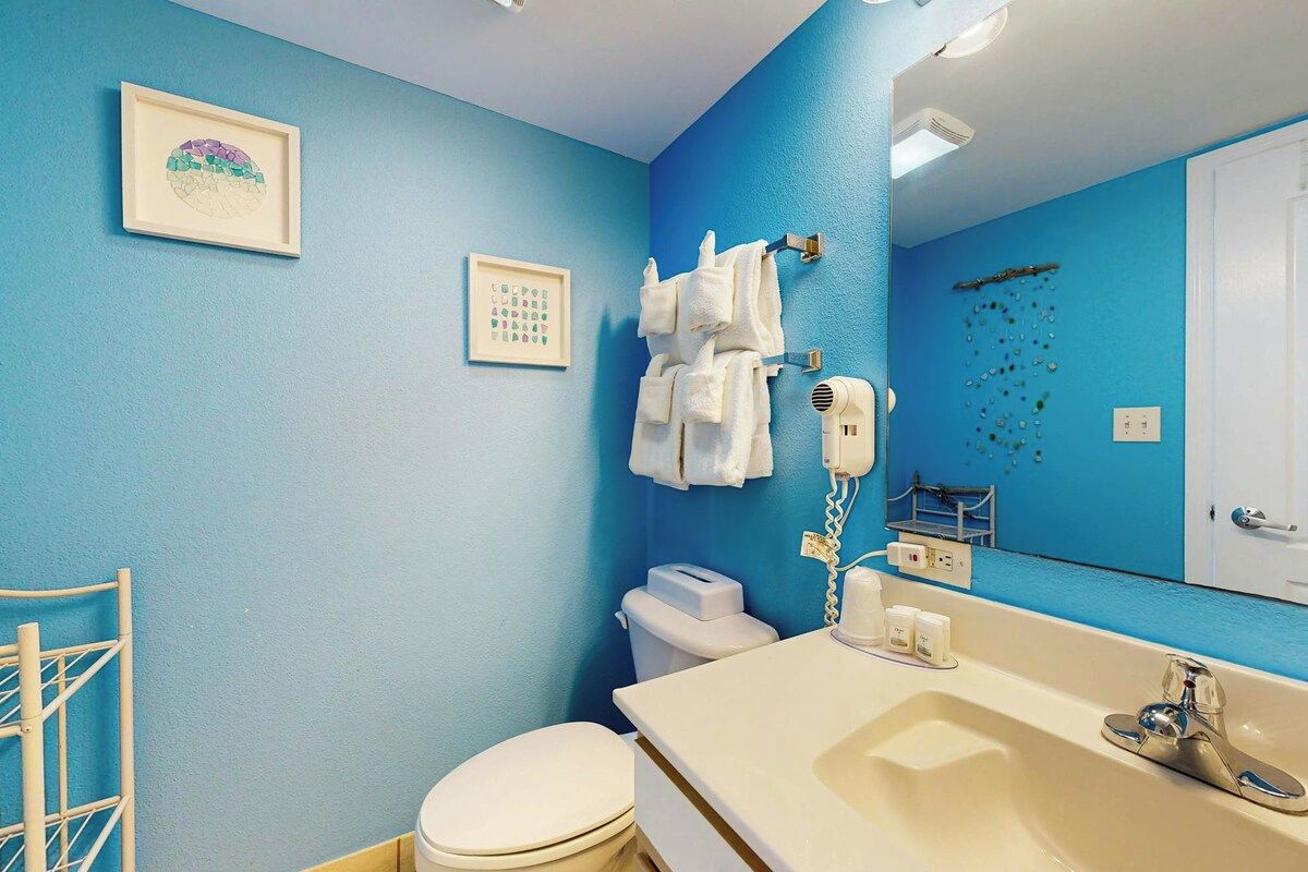Ein Badezimmer mit blauer Wand, Toilette in Weiß, Waschbecken inklusive Spiegel und Handtuchhalter mit Handtüchern.