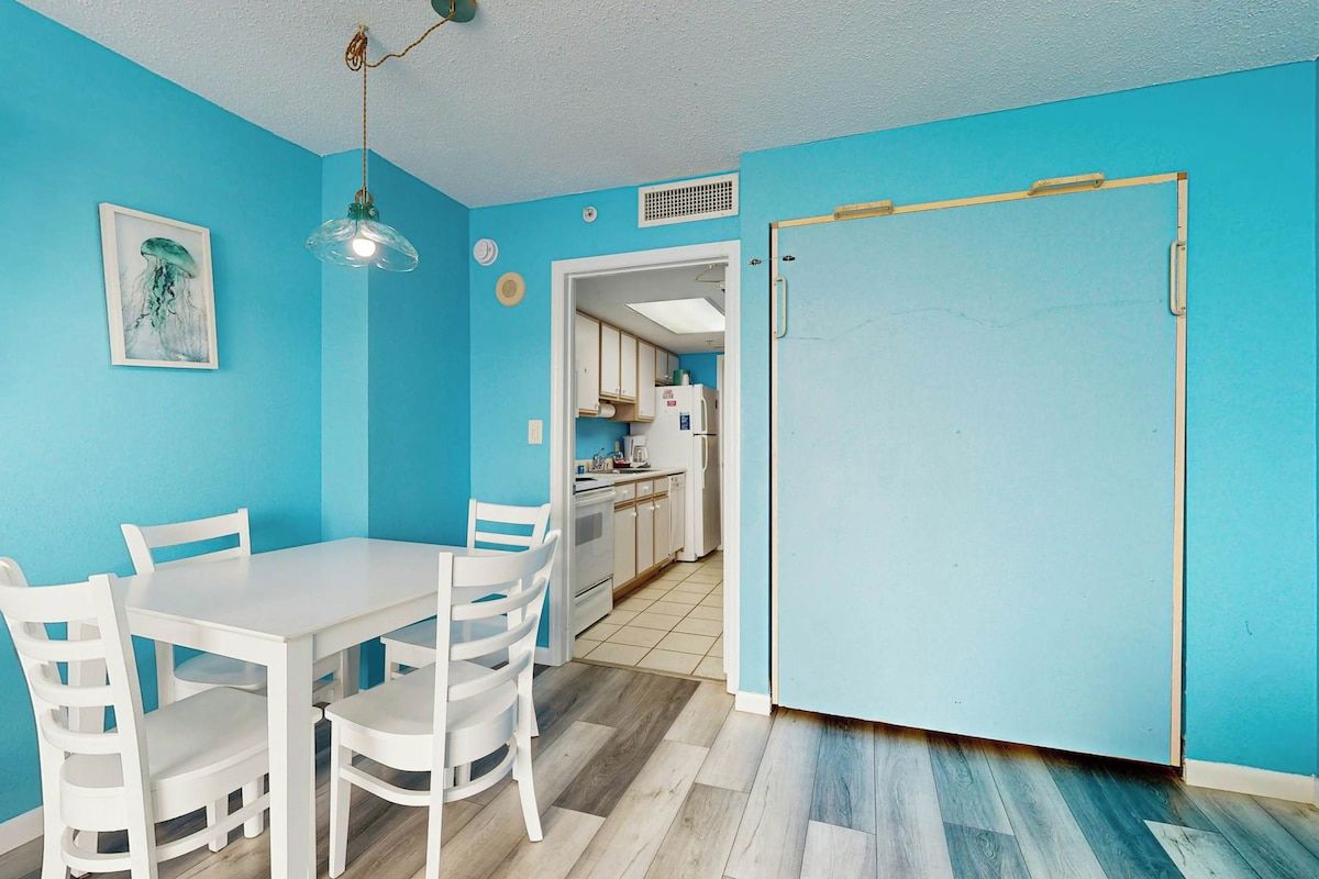 Una habitación con pared azul, una mesa blanca con cuatro sillas, una lámpara colgante y una puerta que da a una zona de cocina.