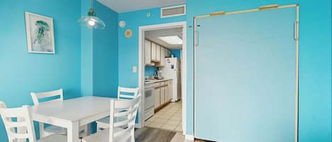 Una habitación con pared azul, una mesa blanca con cuatro sillas, una lámpara colgante y una puerta que da a una zona de cocina.
