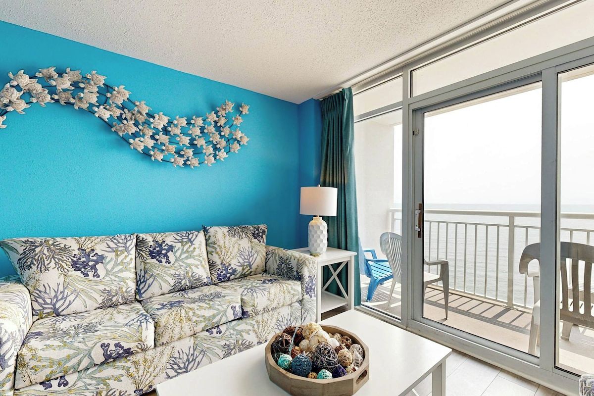 Ein Zimmer mit blauer Wand, einem blumigen Sofa, einer Lampe, einem Tisch mit dekorativen Eiern und Blick auf das Meer durch eine Schiebeglastür.