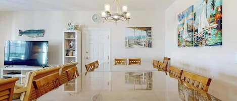 A dining area with a long table, chairs, a chandelier, a television, and artwork on the walls.