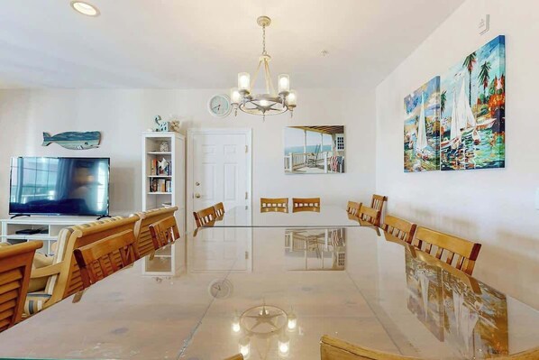 A dining area with a long table, chairs, a chandelier, a television, and artwork on the walls. - Coconut Grove Unit 202 (North Myrtle Beach)