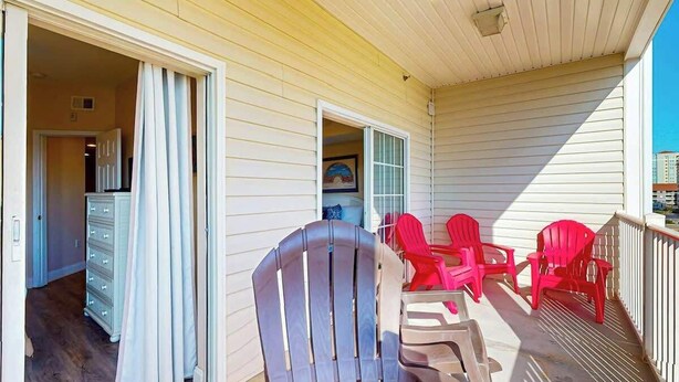 A covered porch with a sliding glass door leading to a room, red Adirondack chairs, and a white dresser. - Coconut Grove Unit 202 (North Myrtle Beach)