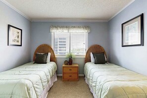 A bedroom with two single beds, a nightstand with a plant, and a window with blinds. - Tilghman Beach & Racquet 214 (North Myrtle Beach)