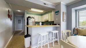 A compact kitchen with white cabinets, a central island with stools, and a dining area with a round table and chairs. - Tilghman Beach & Racquet 214 (North Myrtle Beach)