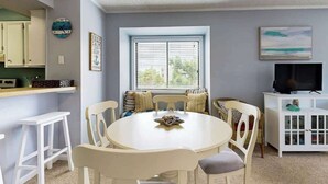A dining area with a round table and chairs, a window with blinds, a television on a cabinet, and a kitchen area with cabinets and a clock. - Tilghman Beach & Racquet 214 (North Myrtle Beach)
