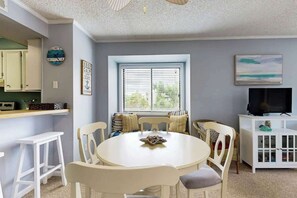 A dining area with a round table and chairs, a window with blinds, a television on a cabinet, and a kitchen area with cabinets and a clock. - Tilghman Beach & Racquet 214 (North Myrtle Beach)