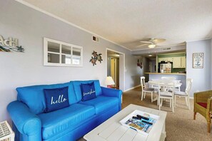 A living room with a blue sofa, a coffee table with magazines, a dining area with white chairs, and a kitchen in the background. - Tilghman Beach & Racquet 214 (North Myrtle Beach)