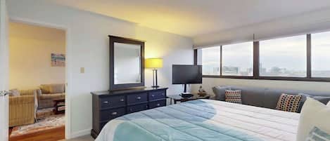 A hotel room with a large bed, a television, a desk with a lamp, and a city view through the windows.