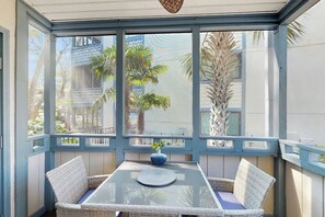 A conservatory with a glass table and wicker chairs, a blue vase, and views of palm trees and buildings. - Summertree Village D-4 (Myrtle Beach)