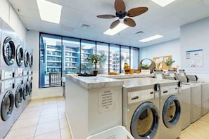 A laundry room with multiple washing machines, a control panel, and a sign indicating room hours. - Renaissance Tower 1114 (Myrtle Beach)
