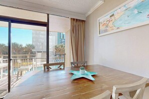 Uma mesa de jantar de madeira com cadeiras, um jogo americano em formato de estrela do mar e uma pequena planta. Uma grande janela oferece vista para a varanda e edifícios externos.