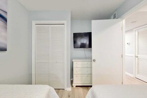 Une chambre avec deux lits, un téléviseur fixé au mur, une commode blanche et un placard avec des portes à persiennes.