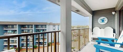 A balcony with a view of a parking lot and a building with multiple floors. There are two white Adirondack chairs on the balcony.