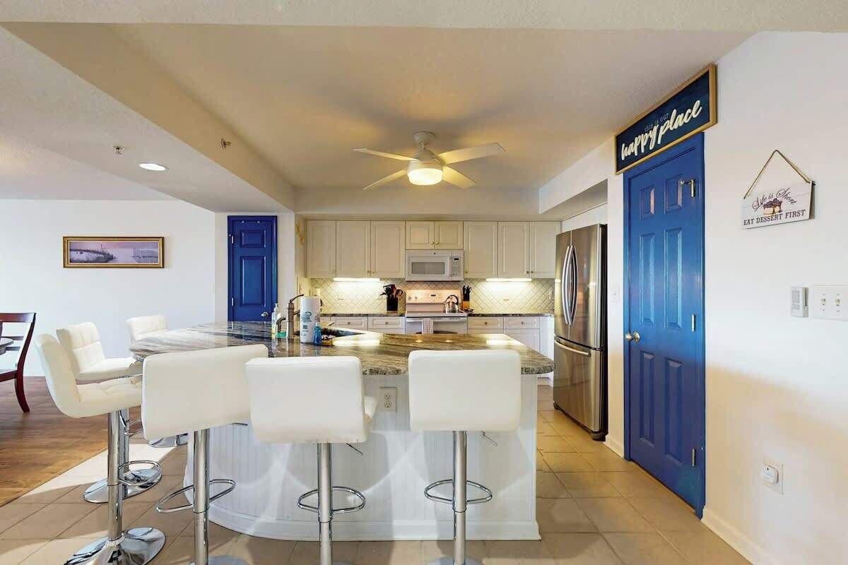 A modern kitchen with white cabinets, a central island with bar stools, a ceiling fan, and a blue door with a sign.