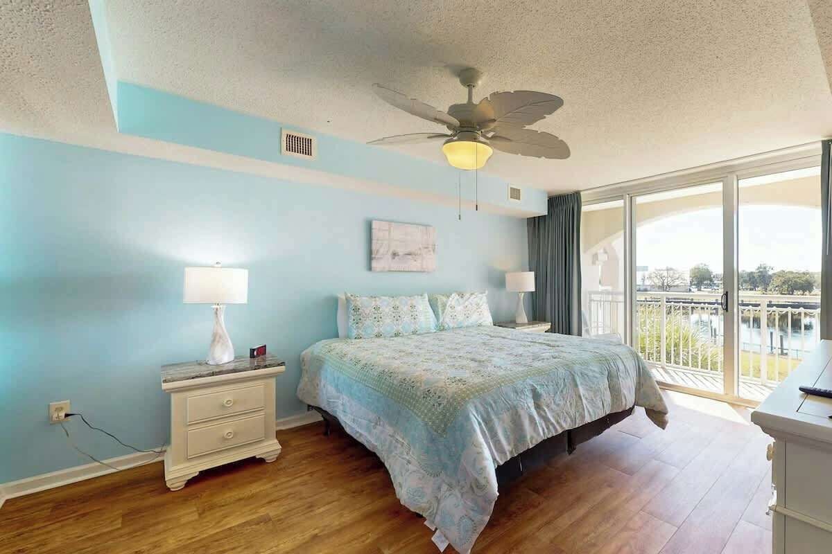 Un dormitorio con cama, mesita de noche, lámpara, ventilador de techo y un amplio ventanal con vistas.