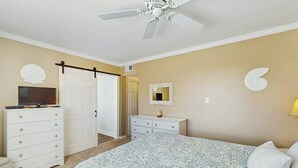 A bedroom with a bed, a dresser, a television, a ceiling fan, and a door leading to another room. - Ocean Bridge Unit 4-B19 (Myrtle Beach)