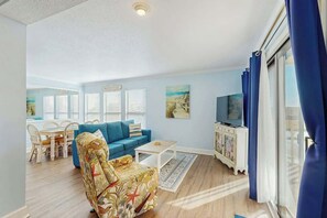 A living room with a sofa, armchair, dining table, and chairs. There is a television and a cabinet with artwork. - Ocean Bridge Unit 4-B19 (Myrtle Beach)