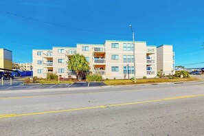 A multi-story residential building with balconies, a parking lot, and a clear sky. - Ocean Bridge Unit 4-B19 (Myrtle Beach)