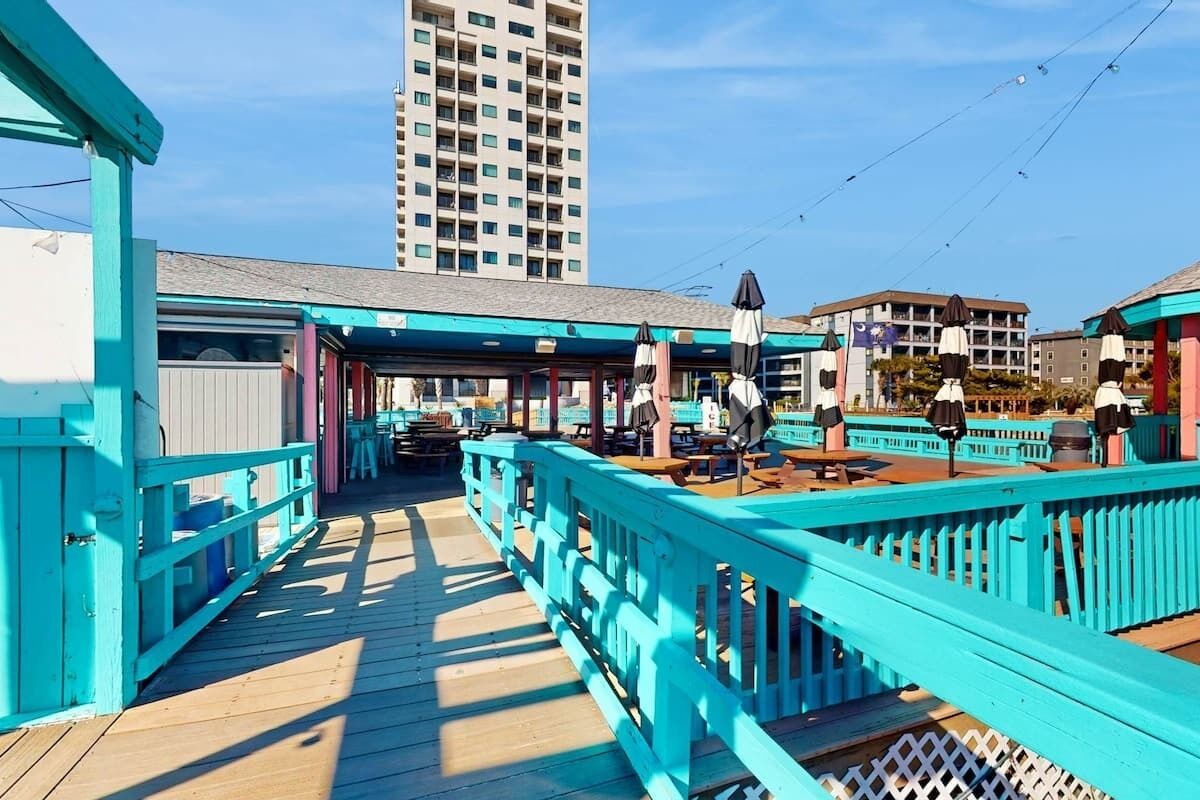 A wooden pier with turquoise railings and picnic tables, leading to a building with a flat roof.