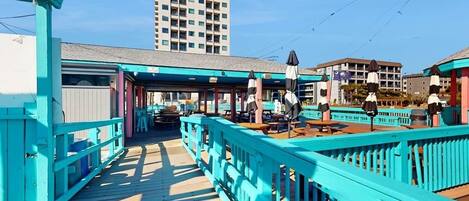 A wooden pier with turquoise railings and picnic tables, leading to a building with a flat roof.