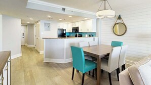 A modern kitchen with white cabinets, a central island, and a dining area with teal chairs. - Grande Cayman Unit 1057 (Myrtle Beach)