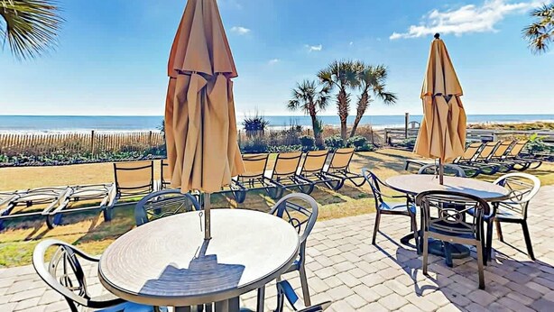 A patio area with round tables and chairs, two umbrellas, and a view of the ocean. - Grande Cayman Unit 1057 (Myrtle Beach)