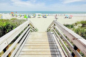 Ein Holzsteg, der zu einem Strand mit Sonnenschirmen und Liegen führt.