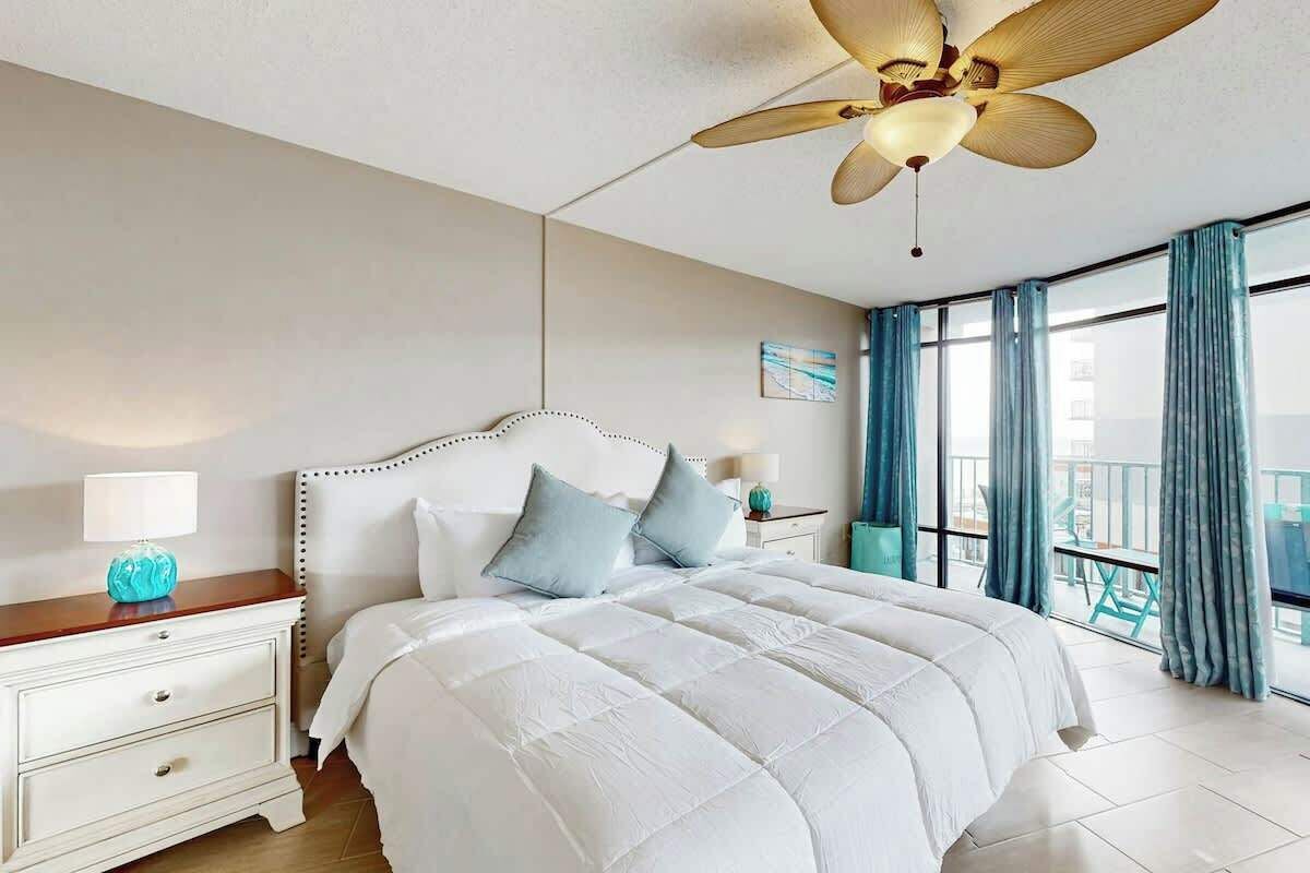 Ein Hotelzimmer mit Bett, Nachttisch, Deckenventilator und einem Balkon mit Vorhängen.