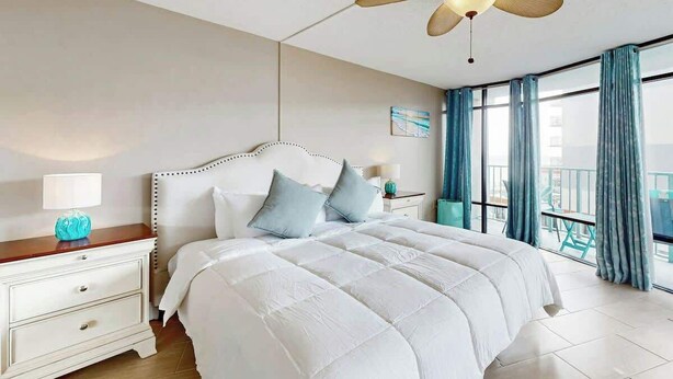 Quarto de hotel com cama, mesa de cabeceira, ventoinha no teto e varanda com cortinas.