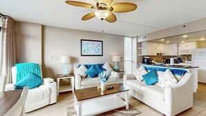 Uma sala de estar moderna com um ventilador de teto, sofás brancos, uma mesa de centro e uma área de cozinha ao fundo.