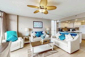 Ein modernes Wohnzimmer mit Deckenventilator, weißen Sofas, einem Couchtisch und einer Küchenzeile im Hintergrund.