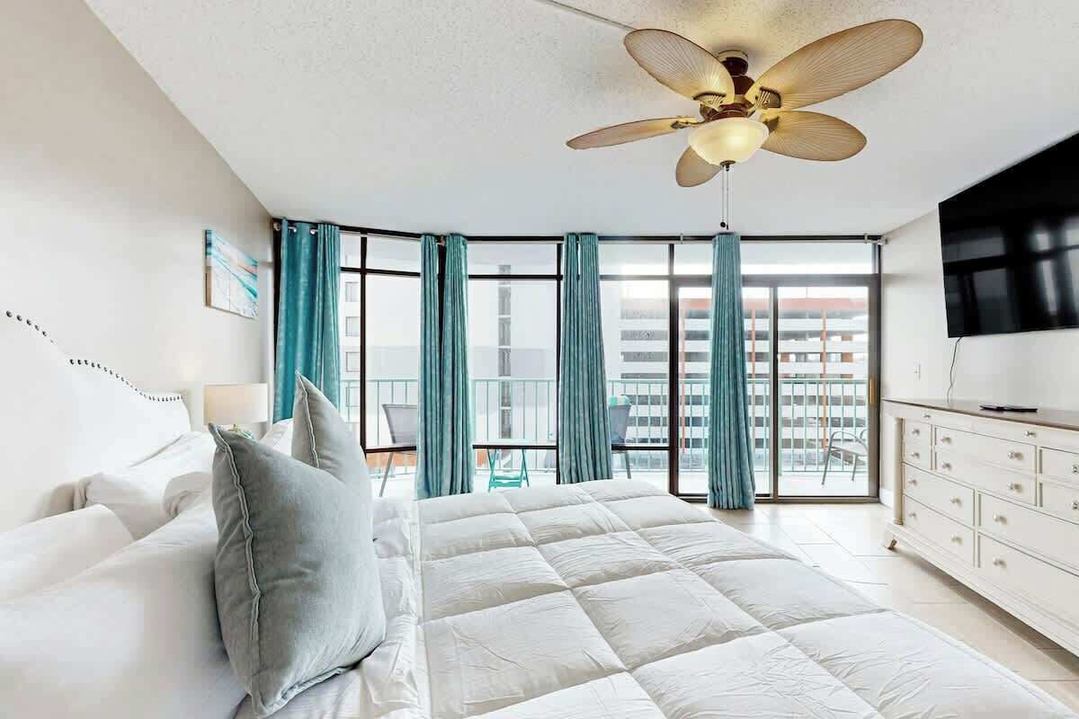 Een hotelkamer met een bed, een plafondventilator, een televisie en een balkon met gordijnen.