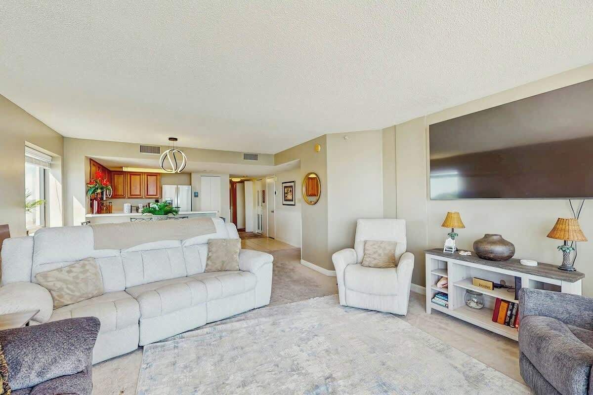 A living room with a sofa, armchairs, a coffee table, and a TV. There is a kitchen in the background.