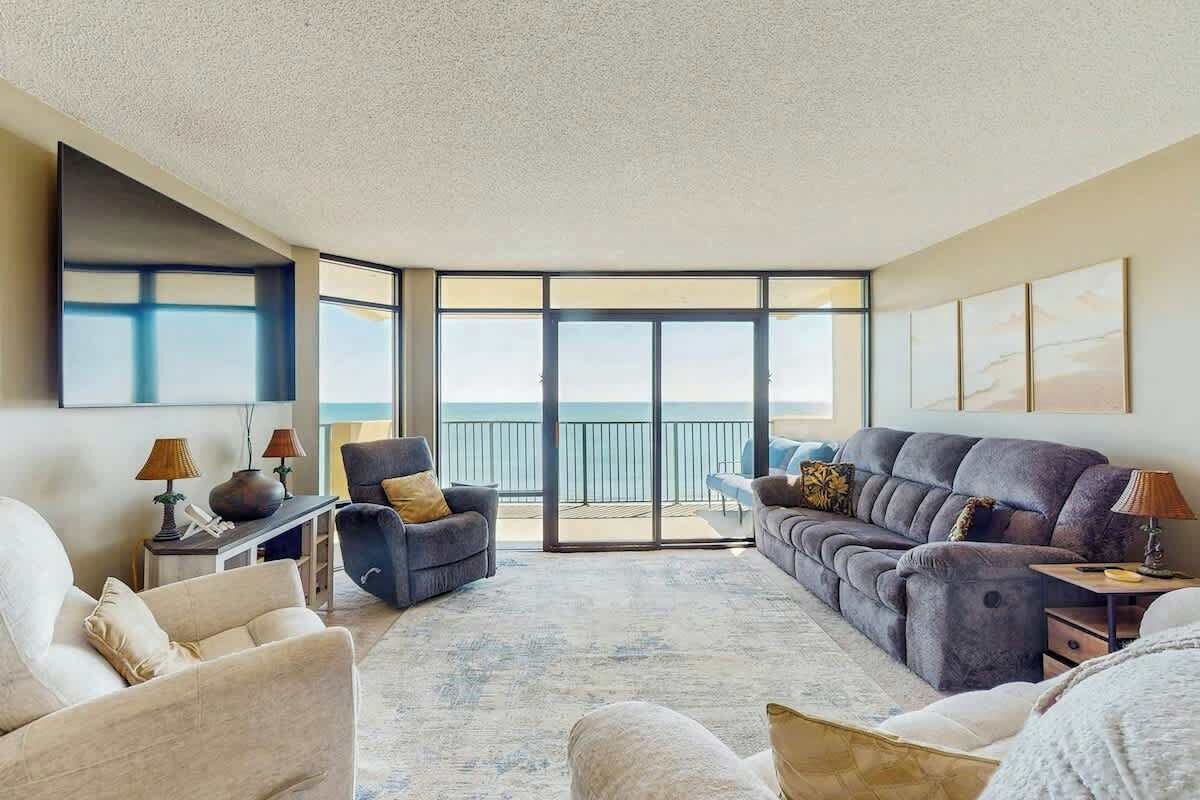 A spacious living area with a large window overlooking a balcony, furnished with a sofa, armchairs, and a coffee table.