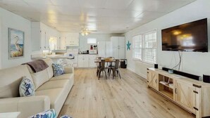 Sala de estar com sofá, mesa de jantar e cadeiras, cozinha com armários e eletrodomésticos, e uma TV de tela plana na parede.