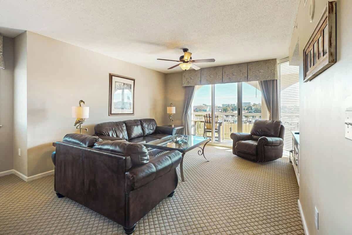 A hotel room with a large window, a ceiling fan, a leather sofa, a glass coffee table, and a view of the city.
