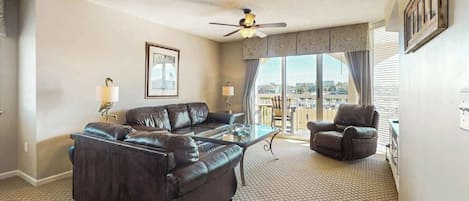 A hotel room with a large window, a ceiling fan, a leather sofa, a glass coffee table, and a view of the city.