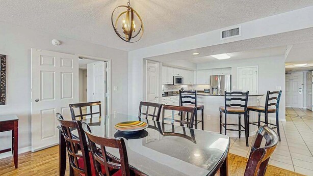 A dining area with a glass table and wooden chairs, a kitchen in the background, and a ceiling light fixture. - Yacht Club Villas 2-304 (North Myrtle Beach)