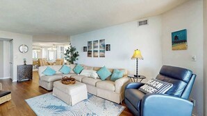 A living room with a sofa, ottoman, and a blue chair. There is a lamp, a rug, and artwork on the walls. - Maisons-sur-Mer  1507 (Myrtle Beach)