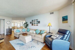 Ein Wohnzimmer mit einer Couch, einem Hocker und einem blauen Sessel. Es gibt eine Lampe, einen Teppich und Kunstwerke an den Wänden.