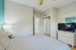 A bedroom with a bed, a television on a dresser, and a door leading to another room. - River Crossing at Barefoot 125 (North Myrtle Beach)